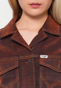 Temno rjav jeans jakna z oranžnim šivom, ki ima klasičen ovratnik, sprednje žepe z poklopcem in vidno oznako Wrangler.