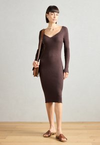 Abito a coste marrone con maniche lunghe e scollo a cuore, indossato con scarpe scivolate marroni, completato da una borsa intrecciata.