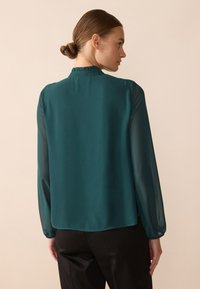 Teal blouse made of lightweight fabric, featuring long sheer sleeves, a ruffled collar, and a relaxed fit. The back is smooth with a slightly curved hem.