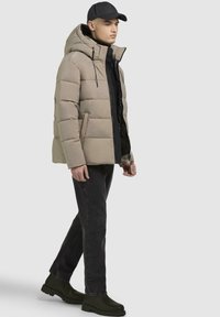 Beige pufferjakke med hætte, lynlåslukning foran og sort inderside. Sort turtleneck, mørke jeans og sorte støvler fuldender outfittet.