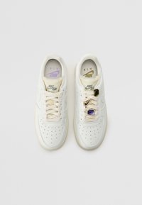 Un par de zapatillas Nike Air Force 1 blancas vistas desde arriba, una con agujetas lisas y la otra adornada con charms tipo gema en las agujetas.