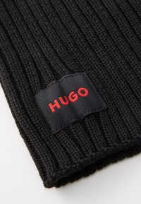 Gorro de punto negro acanalado con una etiqueta de tela rectangular en la que está bordado "HUGO" en rojo. Textura suave y un ajuste ceñido.