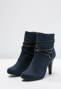 Botas de tobillo de ante azul marino con punta afilada, tacón alto y adornos trenzados alrededor del tobillo; detalladas con un herraje metálico circular.