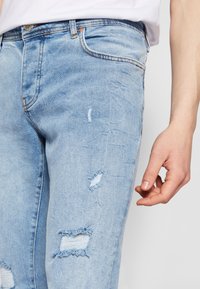 Světle modré džíny z denimu s roztrženými dírami, úzkým střihem a viditelným prošitím. Materiál vypadá měkký s vybledlou strukturou.