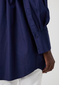 Camicia blu navy con bottoni, maniche lunghe e vestibilità rilassata; presenta un tessuto testurizzato e un orlo leggermente arcuato.