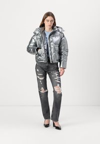 Sølv puffjakke med høy krage, frontlomme og glidelås, kombinert med slitte svarte jeans og svarte høyhælte sko.