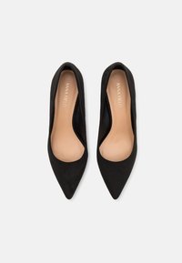 Chaussures plates noires à bout pointu avec semelles beige et marquage "ANNA FIELD", vues de dessus sur fond blanc.