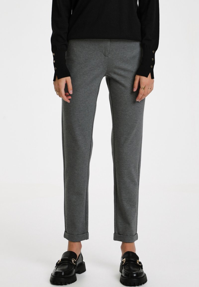 Personne portant un pantalon gris droit avec revers, des mocassins noirs avec boucles dorées, et un haut noir à manches longues avec poignets boutonnés.