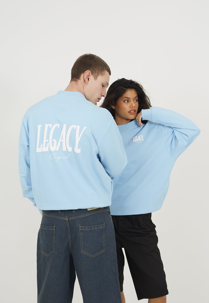 Blå hettegensere med "LEGACY" trykket i hvitt på ryggen og foran. Modell iført mørk denimshorts og en annen i svarte shorts.