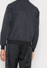 Zwarte nylon bomberjack met geribbelde manchetten en zoom. Heeft een label op de achterkant; een gladde textuur met een licht glanzende afwerking.