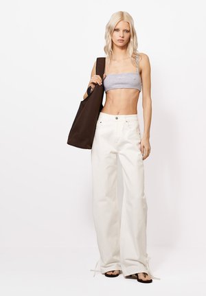 Młoda kobieta o długich blond włosach nosi szary top typu tube, szerokie białe spodnie, czarne sandały i niesie dużą ciemnobrązową torbę na ramię.