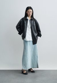 Frau steht mit den Händen in den Taschen, trägt eine schwarze Lederjacke, ein weißes Hemd, einen langen hellblauen Jeansrock und schwarze Absätze.