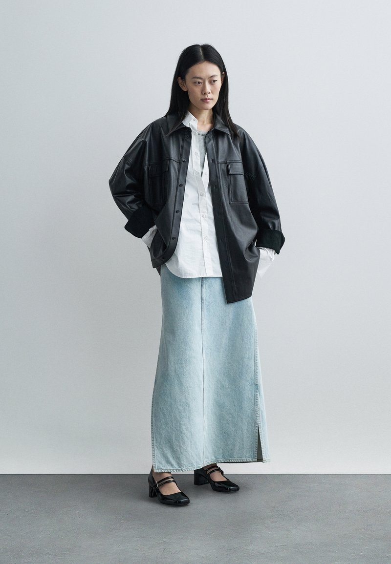 Frau steht mit den Händen in den Taschen, trägt eine schwarze Lederjacke, ein weißes Hemd, einen langen hellblauen Jeansrock und schwarze Absätze.