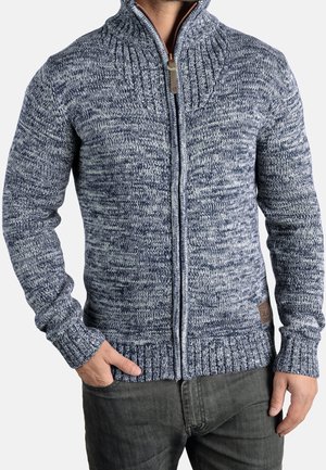 Mann trägt einen blau-weiß melierten Strick-Zippullover mit gerippten Bündchen und Kragen sowie dunkelgraue Hose, eine Hand in der Tasche.