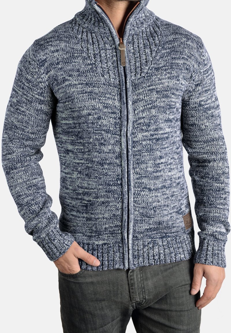 Homme portant un pull zippé en maille chinée bleu et blanc avec poignets et col côtelés, et un pantalon gris foncé, une main dans la poche.