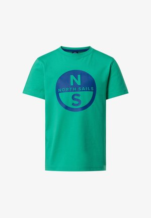 Garçon aux cheveux blonds portant un t-shirt vert "North Sails", un pantalon bleu marine, des baskets blanches, debout les mains dans les poches, souriant.