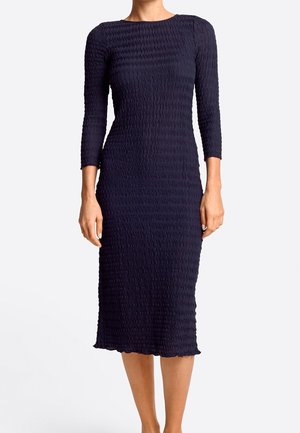 Dopasowana granatowa sukienka midi z długimi rękawami, z teksturowanym falistym wzorem, noszona przez stojącą osobę na jednolitym tle.