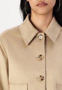 Veste chemise en faux daim beige avec un col évasé, deux poches frontales et de grands boutons ronds avec un design marbré.
