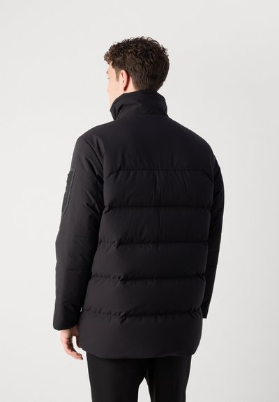 Veste noire matelassée avec un col haut, un quilting horizontal et un patch logo sur la manche gauche. Texture lisse et coupe décontractée.