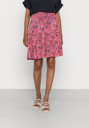 Jupe enveloppante imprimée en rose vif avec des motifs floraux, dotée d'une taille froncée et d'un design en couches. Portée avec des sandales neutres.