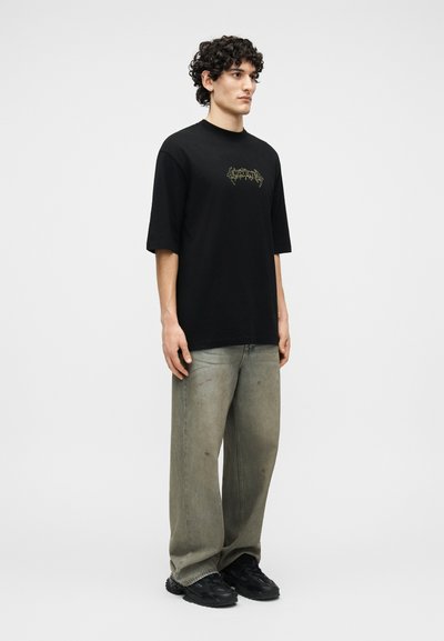 Νέος άνδρας με σγουρά μαλλιά που φοράει μαύρο γραφικό t-shirt, γκρι τζιν με φαρδιά μπατζάκια και ξεθωριασμένα σημεία, και μαύρα αθλητικά παπούτσια, στέκεται μπροστά σε ουδέτερο φόντο.