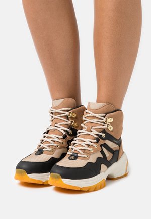Beige and black hiking boots with a padded collar, textured fabric, white rubber sole, and gold hardware accents. Features contrasting elements.