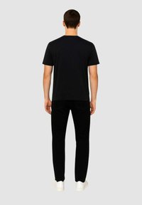 T-shirt nera a maniche corte, in materiale cotone, taglio dritto. Abbinata a pantaloni neri e sneaker bianche, vista da dietro su uno sfondo grigio.