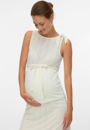 Rase naine kreemikas varrukateta kleidis, millel on seotud vöö, hellalt hoides oma kõhtu ja naeratades silmad kinni.