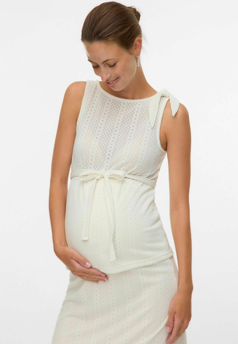 Femme enceinte en robe crème sans manches avec une ceinture nouée, caressant doucement son ventre et souriant les yeux fermés.