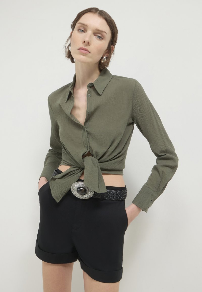 Camicia verde oliva a maniche lunghe con bottoni legata in vita, abbinata a pantaloni corti neri a vita alta e una cintura nera intrecciata con accessori in argento.
