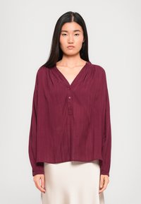 Blouse à manches longues bordeaux avec un col en V et une fermeture à boutons. Présente un tissu souple et texturé avec de légères plis et des poignets élastiqués.