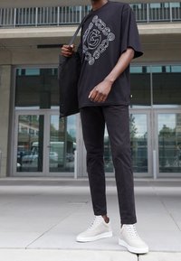 Černé oversized tričko s bílou grafikou, černé slim-fit kalhoty a světle šedé vysoké tenisky. Stojí s černou tote taškou.