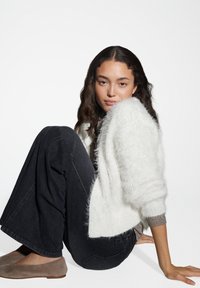 Femme jeune assise, portant un pull blanc douillet, un jean large noir et des ballerines taupe, sur fond blanc uni.