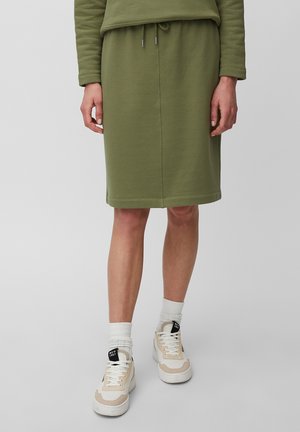 Personne portant une jupe olive longueur genou, un sweat-shirt assorti, des chaussettes blanches, et des baskets beige et blanc sur fond gris.