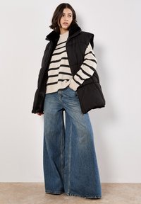 Zwarte puffer vest met een hoge kraag, gecombineerd met een crèmekleurige en zwarte gestreepte trui en een wijdvallende spijkerbroek, die textuur en contrast benadrukt.