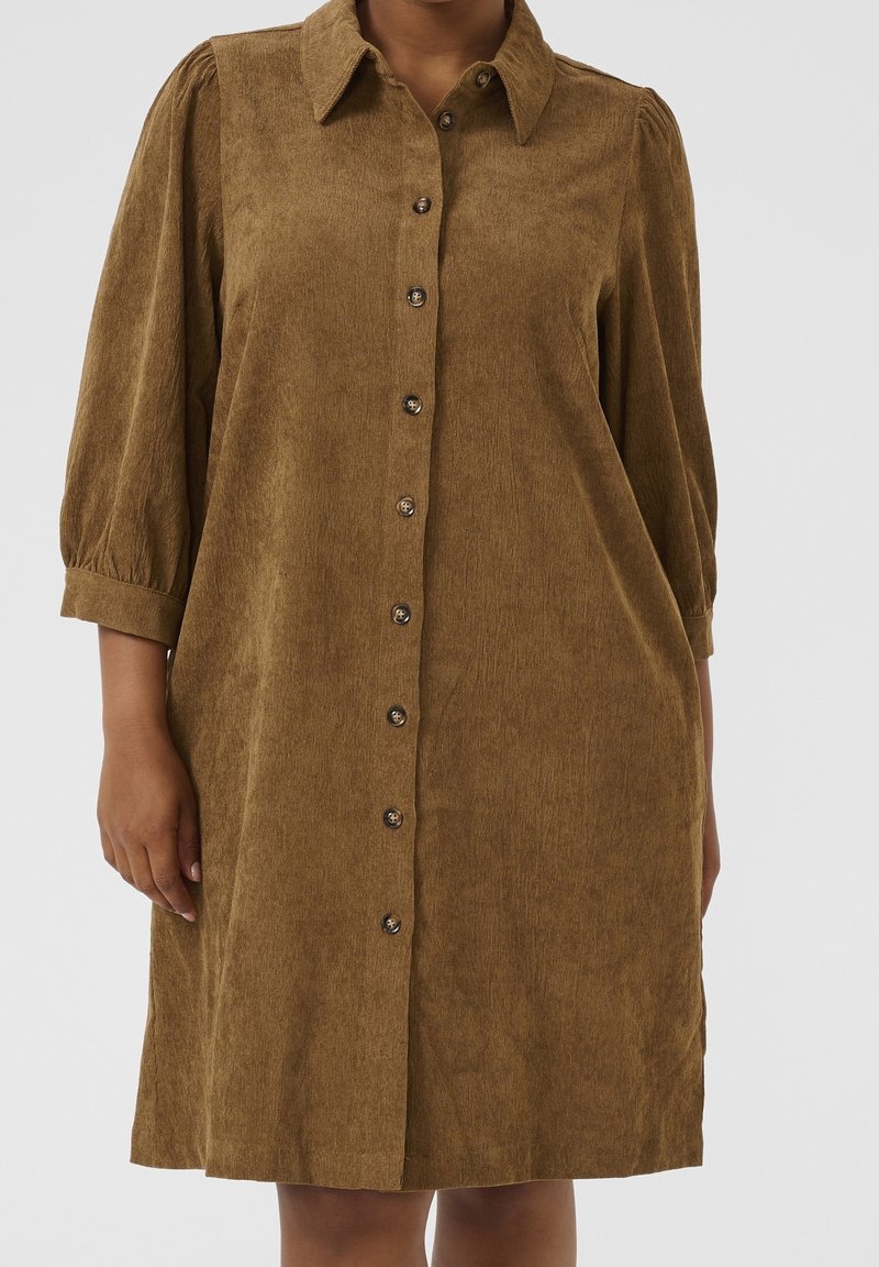 Robe marron en velours côtelé boutonnée, avec manches trois quarts bouffantes et un col, portée par une personne sur un fond uni.