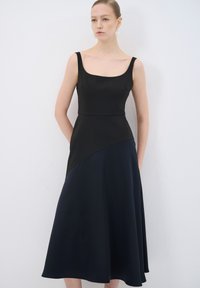 Schwarzes ärmelloses Kleid mit einem taillierten Oberteil und einem schwingenden, kontrastierenden marineblauen Rock. Glatter Stoff mit leichtem Glanz und minimalen Nahtdetails.