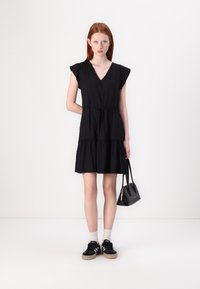 Zwart kortemouwen jurk met een V-hals, geraffineerde taille en gelaagde rok. Gepaarde met zwarte sneakers en een kleine zwarte handtas.