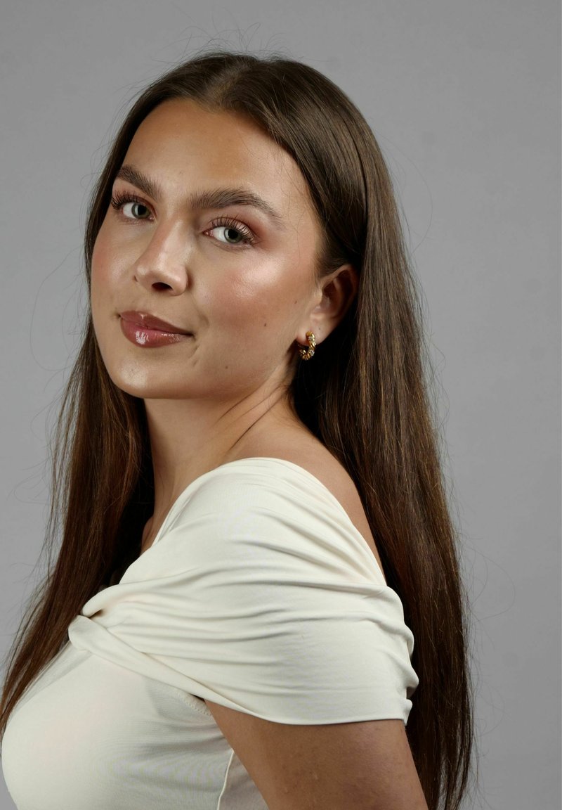 Off-white t-shirt med samlad tygdetalj. Lång, rak, mörk hår. Guldörhängen i form av ringar. Neutral bakgrund.