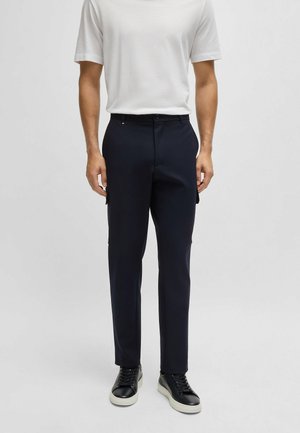 Pantaloni navy su misura in tessuto liscio, con frontale piatto, passanti per cintura e tasche laterali, abbinati a una maglietta bianca a trama e sneakers nere.