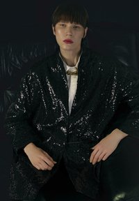 Čierna flitrovaná bunda vo voľnom strihu s golierom a dlhými rukávmi. Nosená cez bielu košeľu, doplnená zlatým retiazkovým náhrdelníkom.