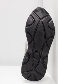 Semelle extérieure en caoutchouc noir avec des motifs de bande de roulement géométriques sur une semelle blanche et grise de sneaker reposant contre une surface blanche.