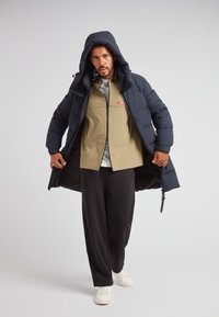 Marineblaue Steppjacke mit Kapuze, offen getragen über einer khakifarbenen Jacke mit Reißverschluss. Locker sitzende schwarze Hose und weiße Turnschuhe vervollständigen das Outfit.