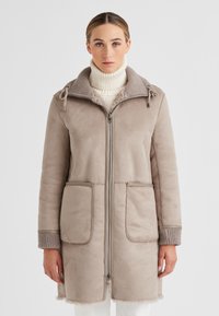 Frau in taupefarbener knielanger Lammfelljacke über einem cremefarbenen Rollkragenpullover, steht vor einem schlichten hellen Hintergrund.