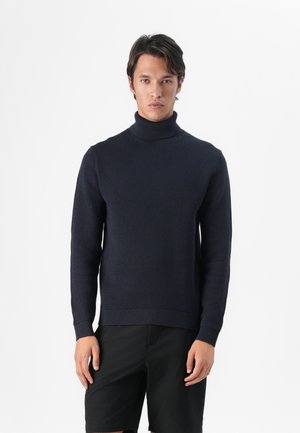 Marineblauwe coltrui gemaakt van ribgebreid materiaal met lange mouwen en een aansluitende pasvorm, gecombineerd met zwarte shorts.