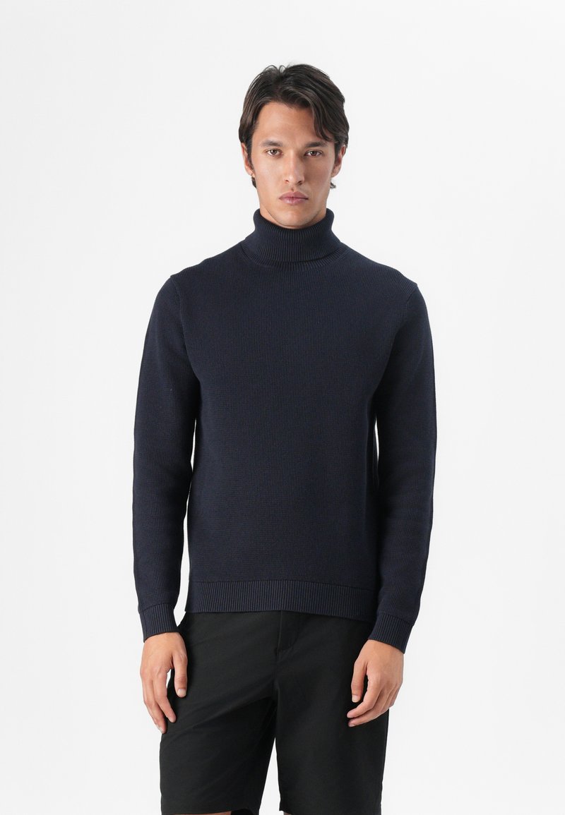 Marineblå turtle-neck genser laget av ribbestrikket materiale med lange ermer og en tettsittende silhuett, kombinert med svarte shorts.