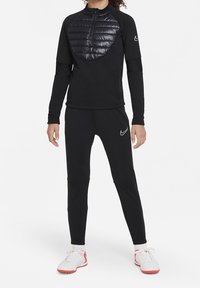 Schwarzes Sportoutfit mit einem langärmligen, mit Reißverschluss versehenen Oberteil, das ein gepolstertes, glänzendes Brustpanel hat, und eng anliegenden schwarzen Hosen, kombiniert mit weißen und roten Schuhen.