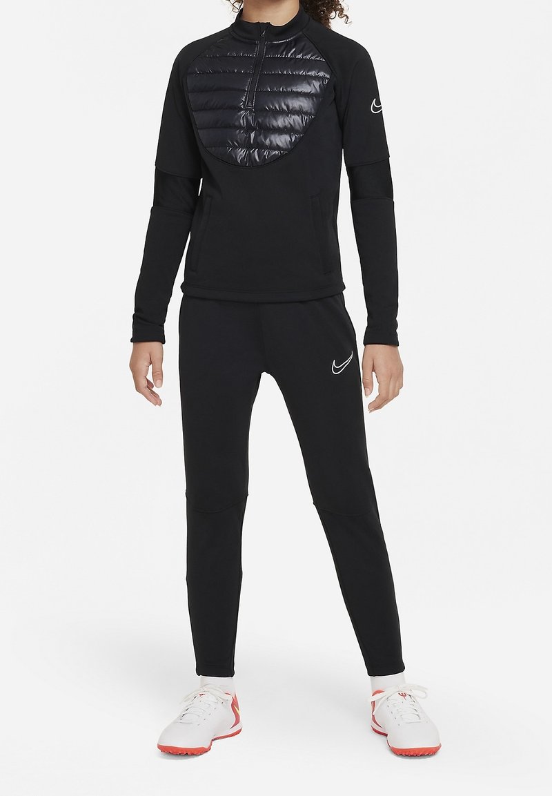 Schwarzes Sportoutfit mit einem langärmligen, mit Reißverschluss versehenen Oberteil, das ein gepolstertes, glänzendes Brustpanel hat, und eng anliegenden schwarzen Hosen, kombiniert mit weißen und roten Schuhen.