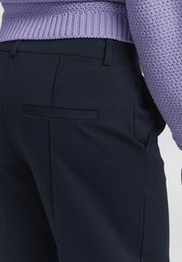 Nærbilde av en person med marineblå skreddersydde bukser med en baklomme og en lavendelstrikket genser.