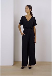 Svart kortärmad jumpsuit med V-ringning, elastisk midja och vida ben. Buren med svarta spetsiga skor på ett trägolv.